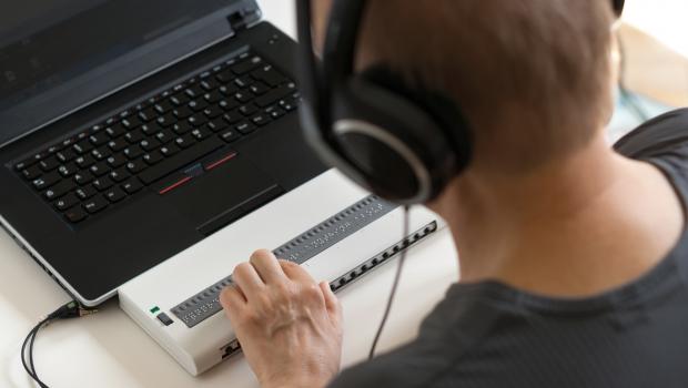 A person using a laptop computer wearing headphones and touching a braille output device.