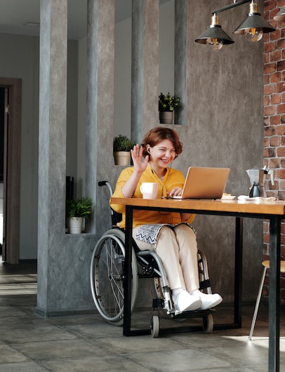 woman in wheelchair sitting at desk with computer, smiling, and waving